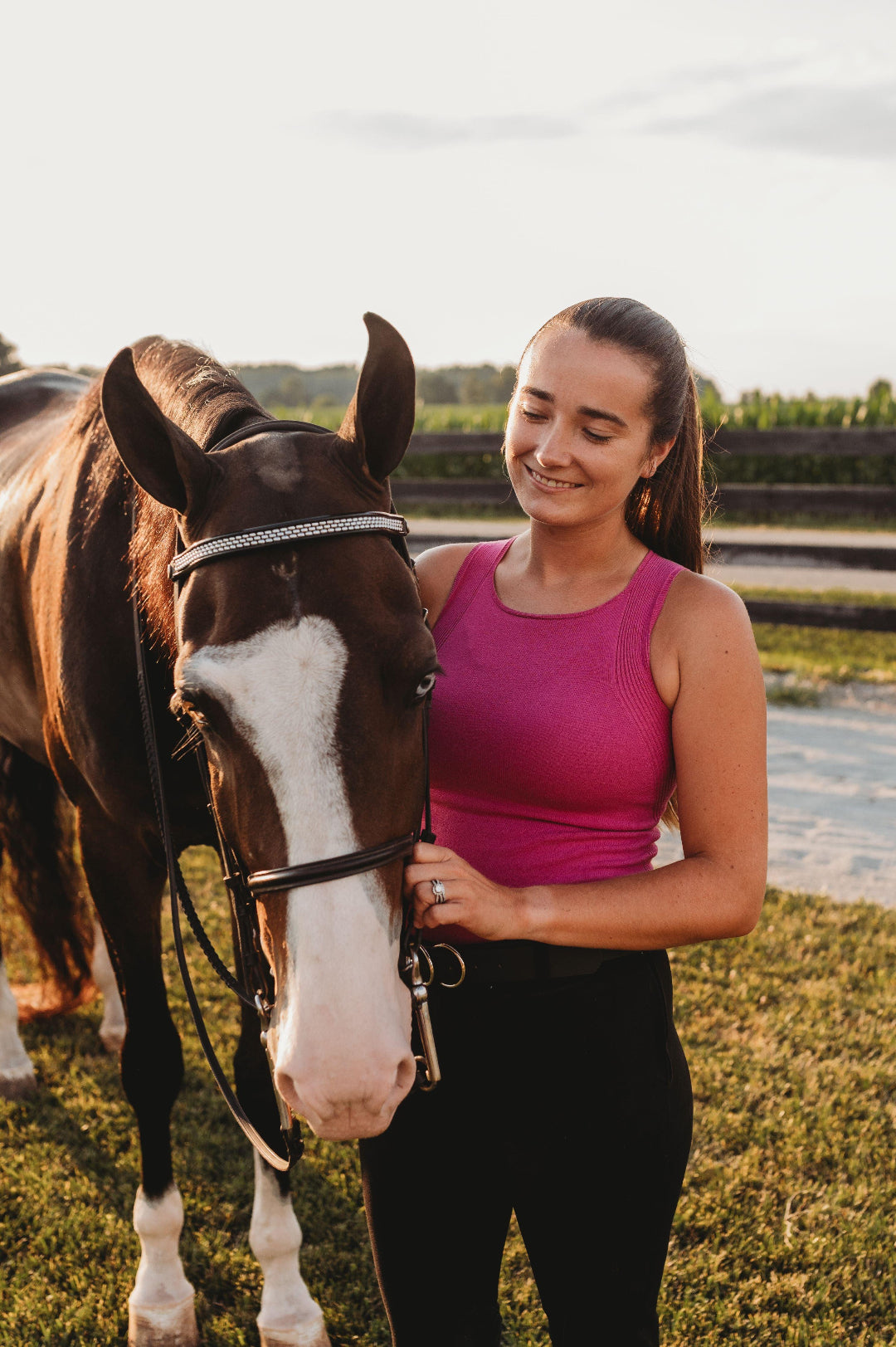 Woman in a pink tank top standing next to a brown horse outdoors.